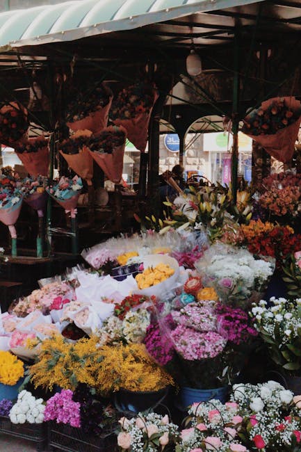 A vibrant display of fresh floral bouquets at an outdoor market stall, featuring a variety of flowers including pink roses, white lilies, yellow mimosa, and purple statice, arranged in large baskets a