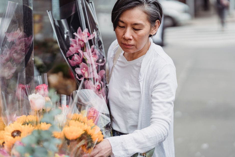 A woman with short, dark grey hair dressed in a white top and light cardigan is selecting a floral arrangement at an outdoor flower shop. She is holding a bouquet of pink orchids wrapped in clear plas
