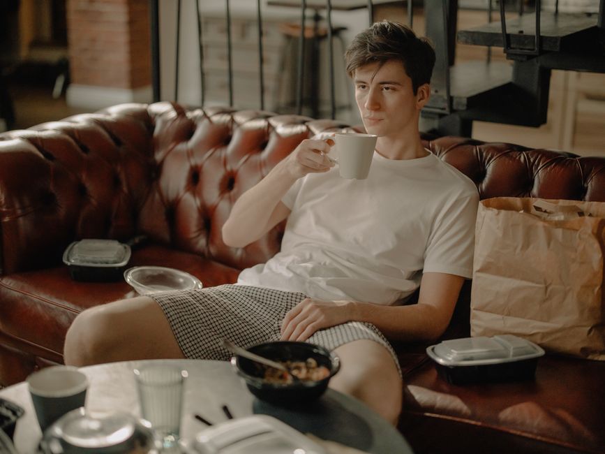 A young man with dark, tousled hair is sitting on a brown leather Chesterfield sofa in a cozy, modern cafe setting illuminated by warm ambient lighting. He is holding a white ceramic mug with his righ