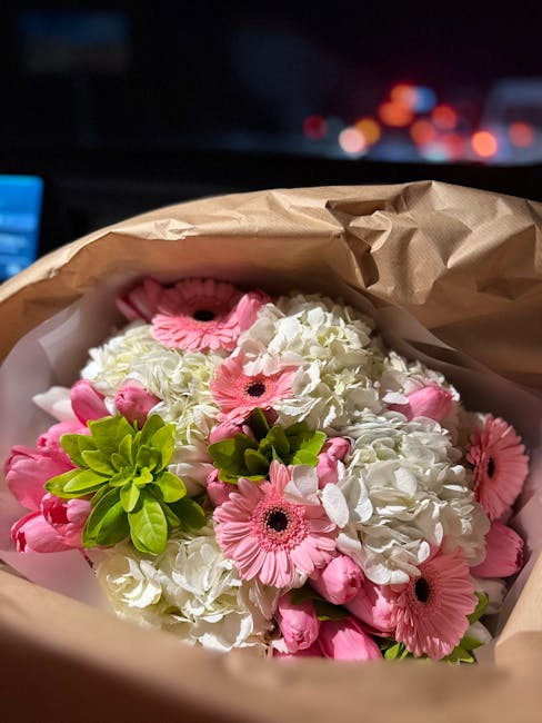 A bouquet of fresh flowers, prominently featuring light pink gerberas with dark centers, white hydrangeas, and bright green foliage, all arranged in a compact, rounded style. The flowers are wrapped i