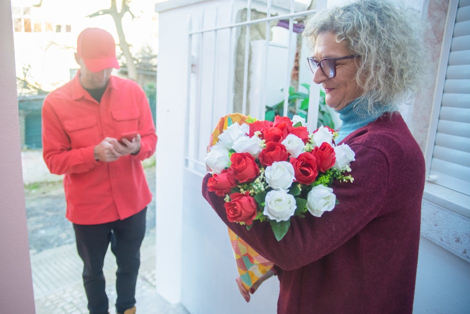 A woman with curly gray hair, wearing glasses and a burgundy cardigan over a light blue turtleneck, holds a floral bouquet featuring red and white roses wrapped in colorful paper, standing outdoors ne
